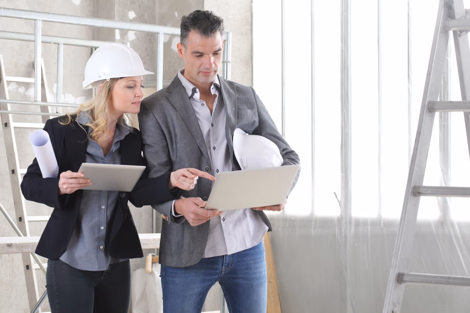 Two construction professionals wearing hard hats reviewing plans on a tablet and laptop at a construction site.