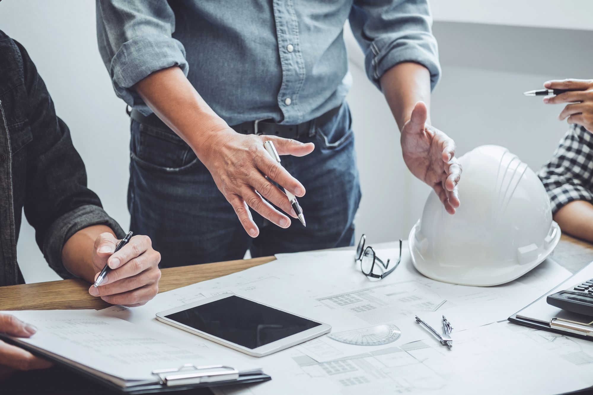 People discussing architectural plans with a tablet, clipboard, white hard hat, and drafting tools on a table.