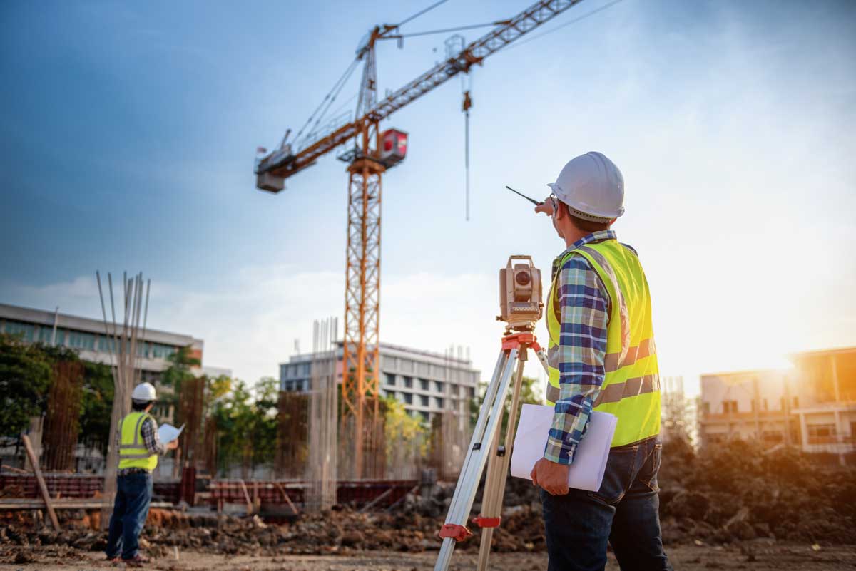 Construction worker in a safety vest and hard hat using surveying equipment at a building site with a crane and another worker in the background.