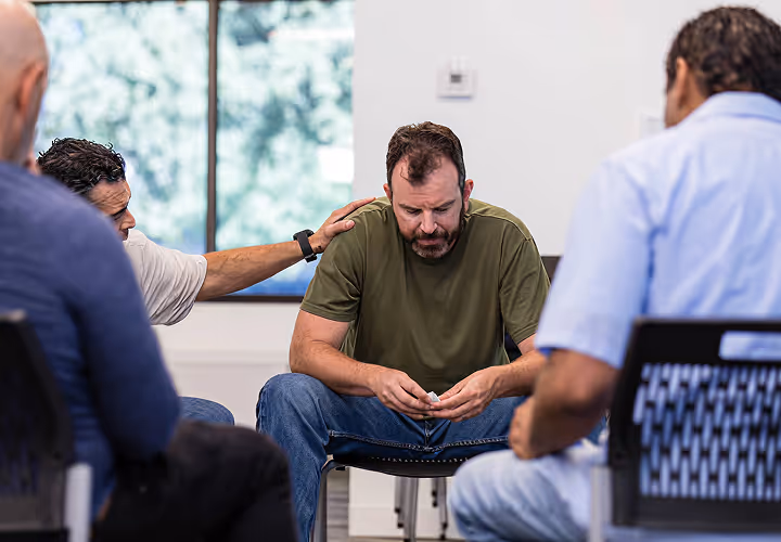 Group therapy session with a man in a green shirt looking down, being comforted by another man.