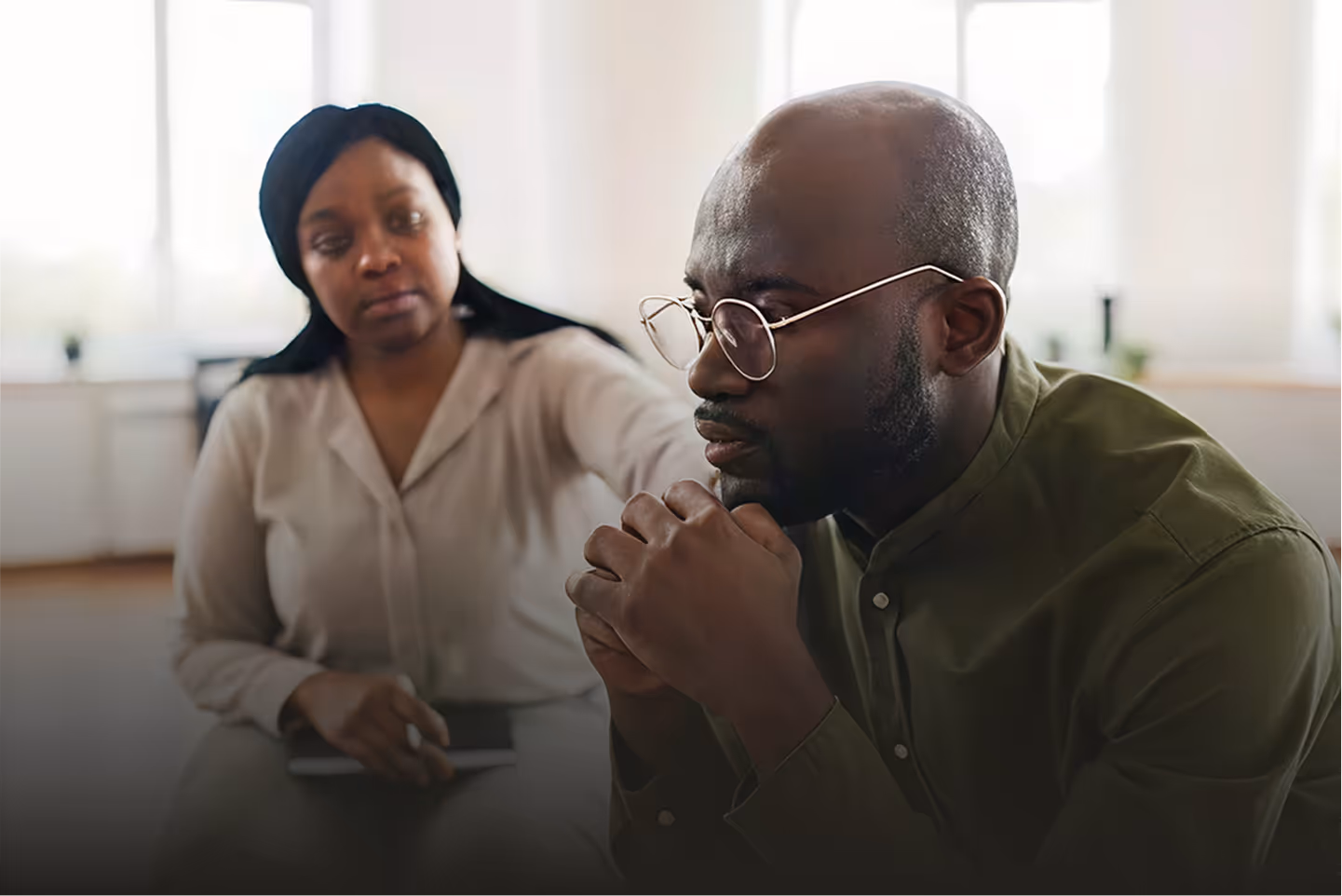 Concerned woman comforts a thoughtful man wearing glasses sitting indoors in a well-lit room.