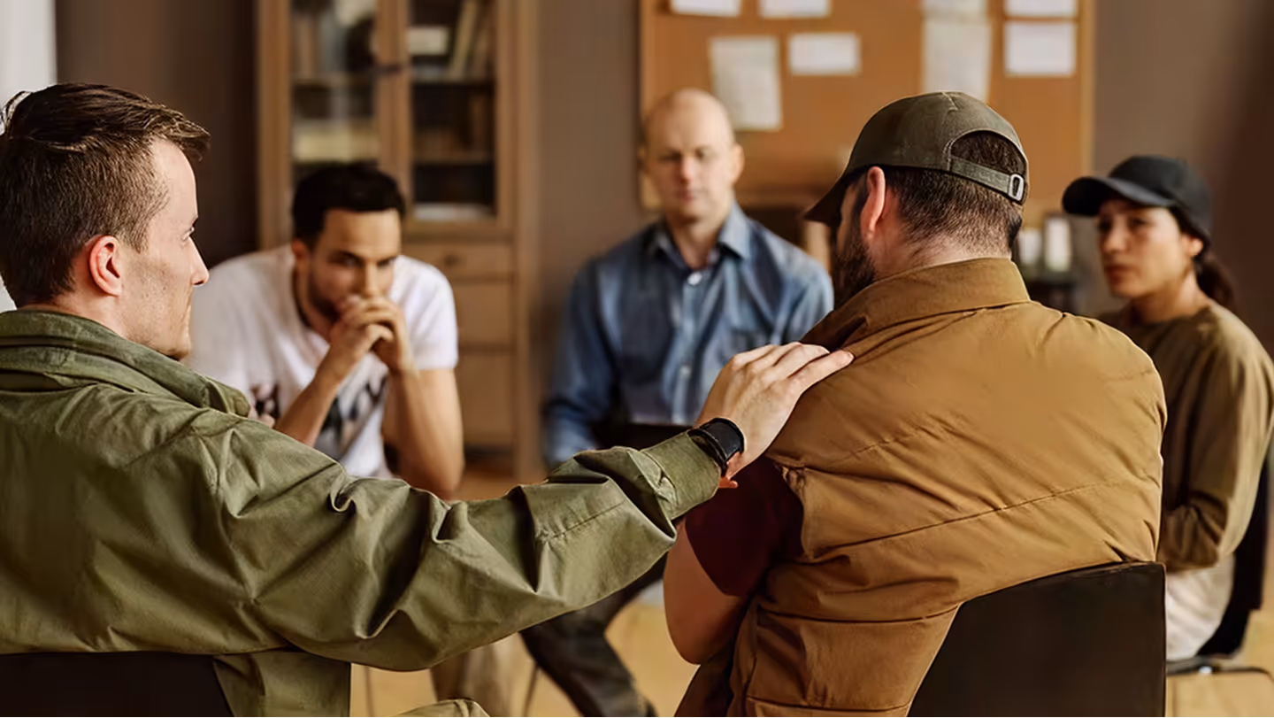 Group therapy session with five adults sitting in a circle, one person comforting another by placing a hand on their shoulder.