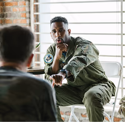 A man in military uniform sitting and pointing while speaking to another person.