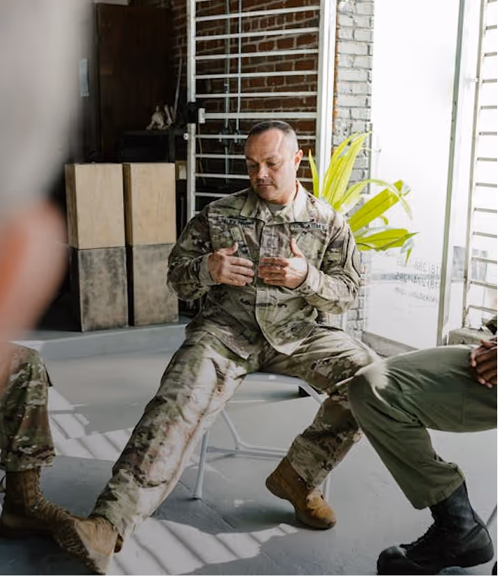 A man in military camouflage uniform sitting on a chair with legs stretched out, talking to others partially visible.