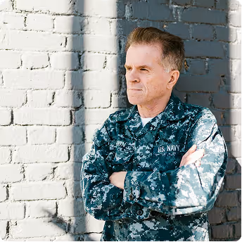 Man in US Navy camouflage uniform standing with arms crossed against a brick wall with sunlight and shadow.