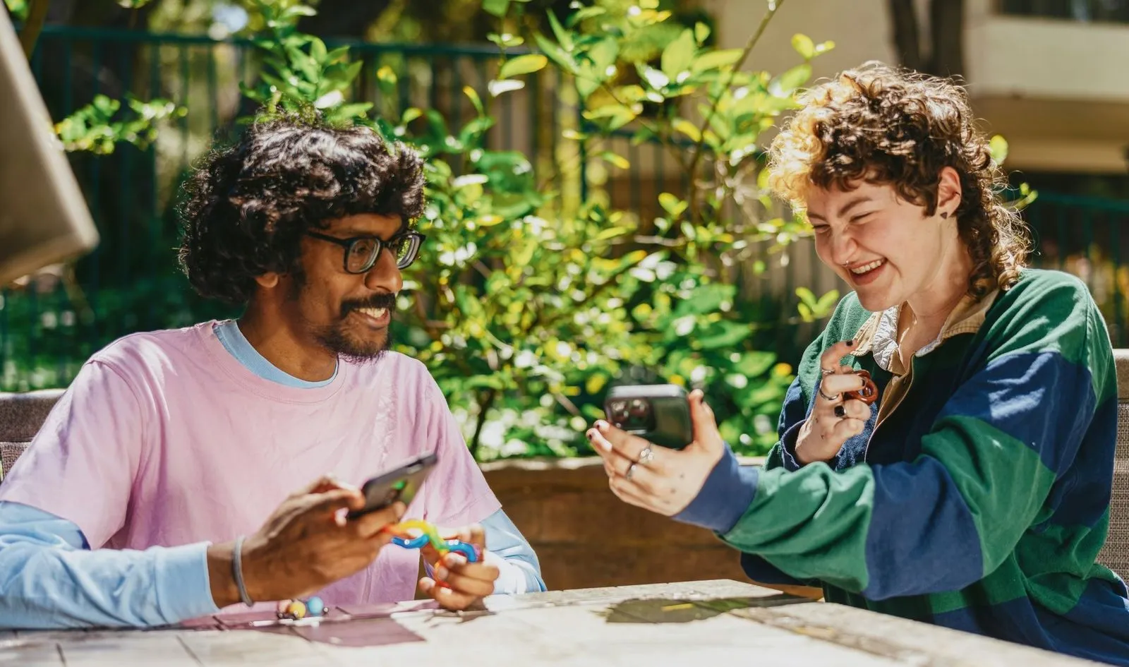 Two people sit outdoors at a table amidst lush greenery, smiling and looking at their phones. The scene conveys a joyful and relaxed atmosphere.