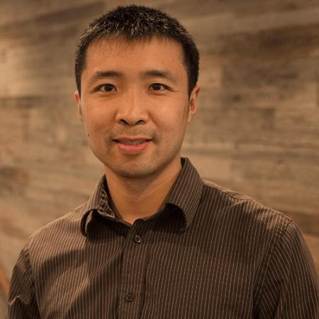 Portrait of a smiling man with short black hair wearing a dark striped button-up shirt against a blurred wooden background.