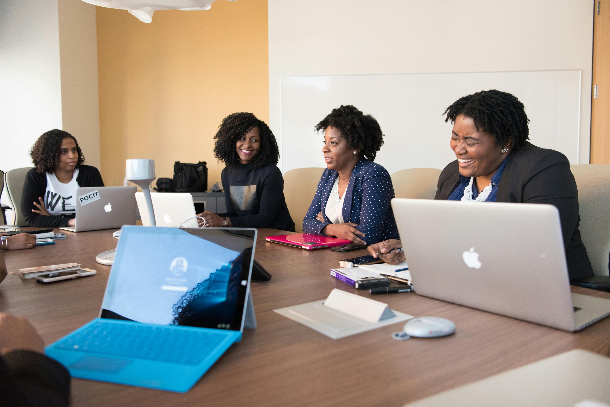 Four women with laptops sitting around a conference table engaged in a meeting, one woman is smiling and another is laughing.