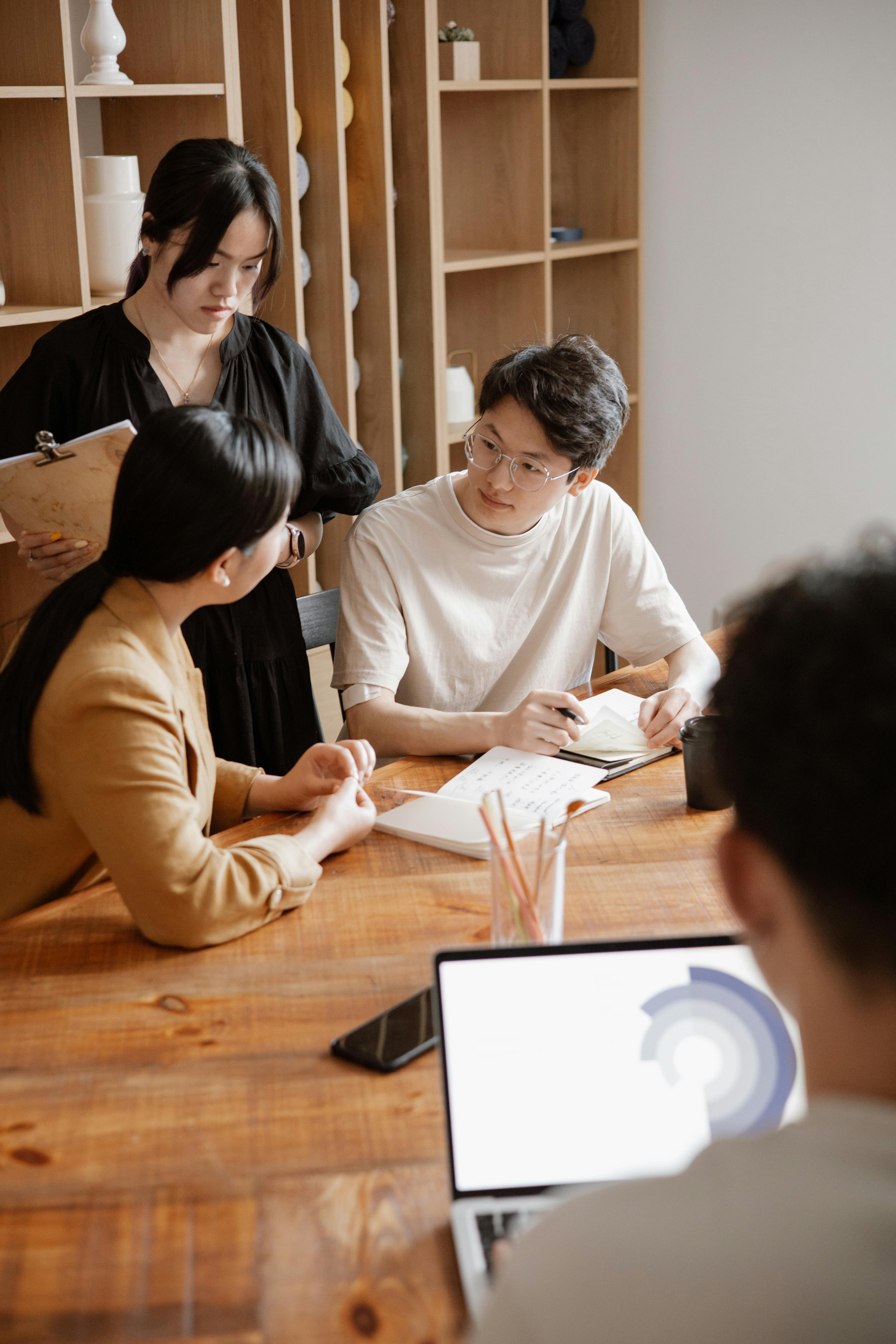 Three people engaged in discussion around a wooden table with notebooks and a laptop, in a modern office setting.