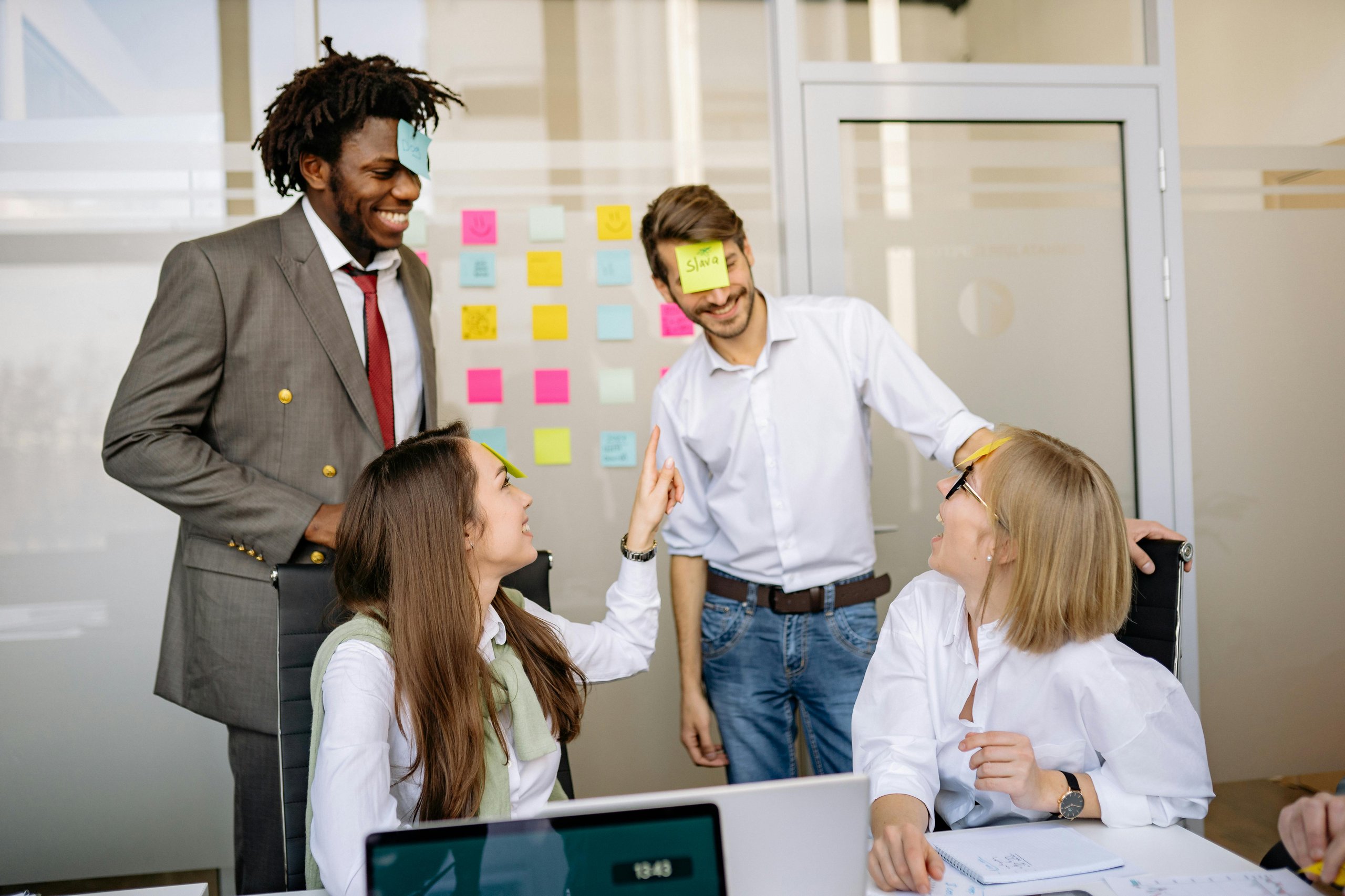 Four coworkers smiling and playing with sticky notes on their foreheads in a bright office.