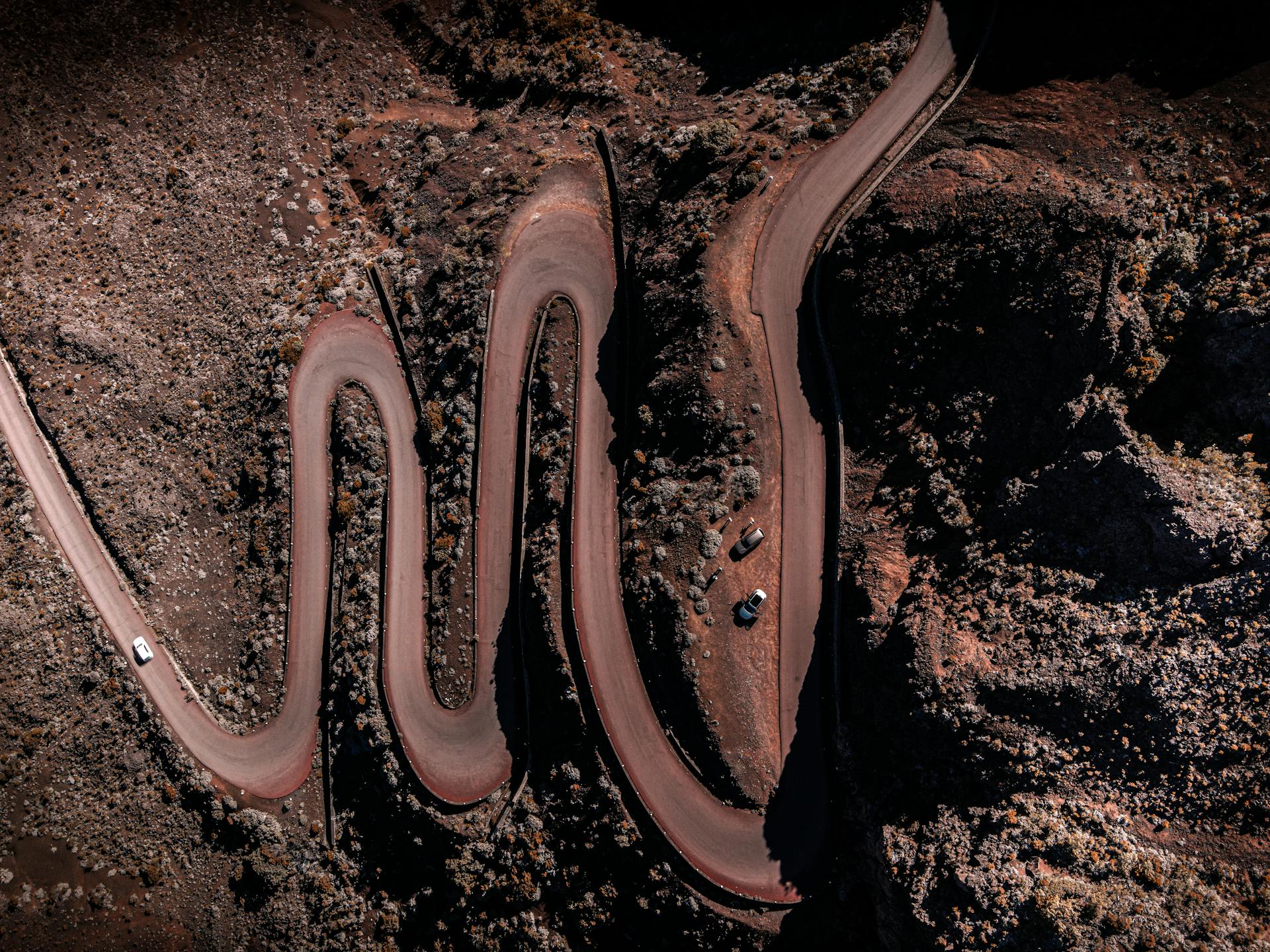 Aerial view of a winding mountain road with sharp hairpin turns and three parked cars on a central curve.