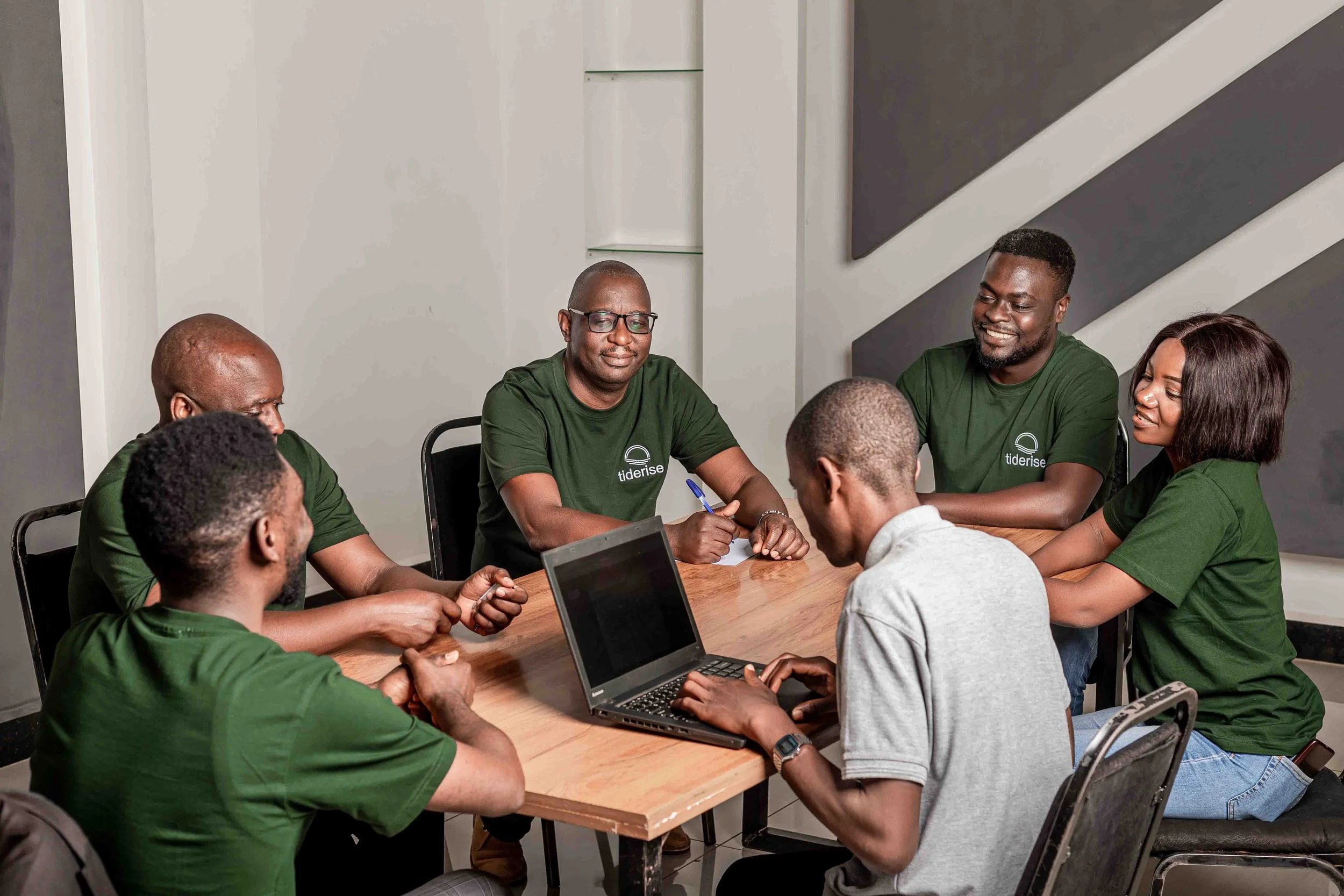 Six people sitting around a wooden table, five wearing green TideRise t-shirts, engaging in a group discussion with one person using a laptop.