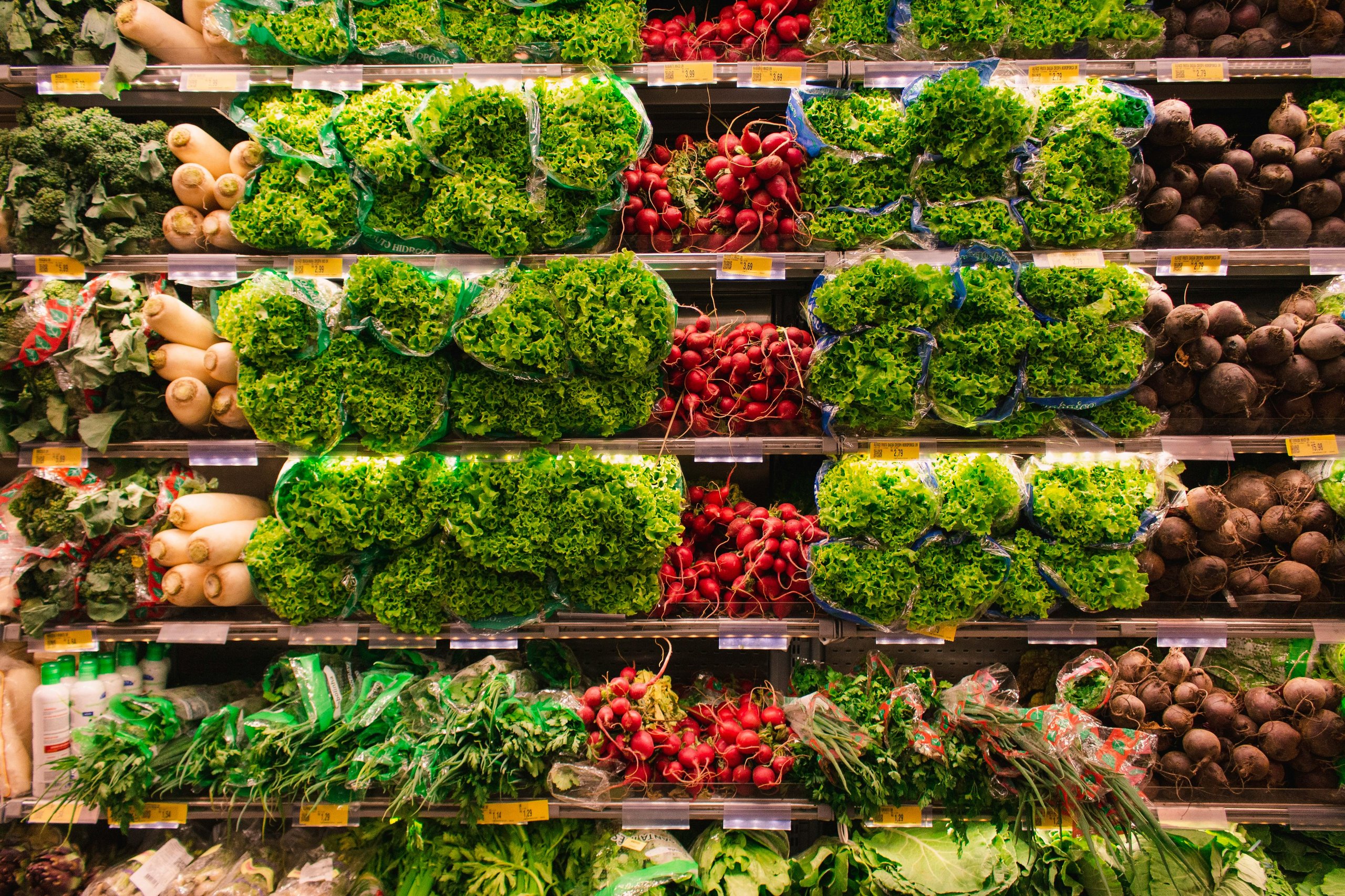 Supermarket produce shelf stocked with bags of leafy green lettuce, bunches of red radishes, white radishes, and beets.