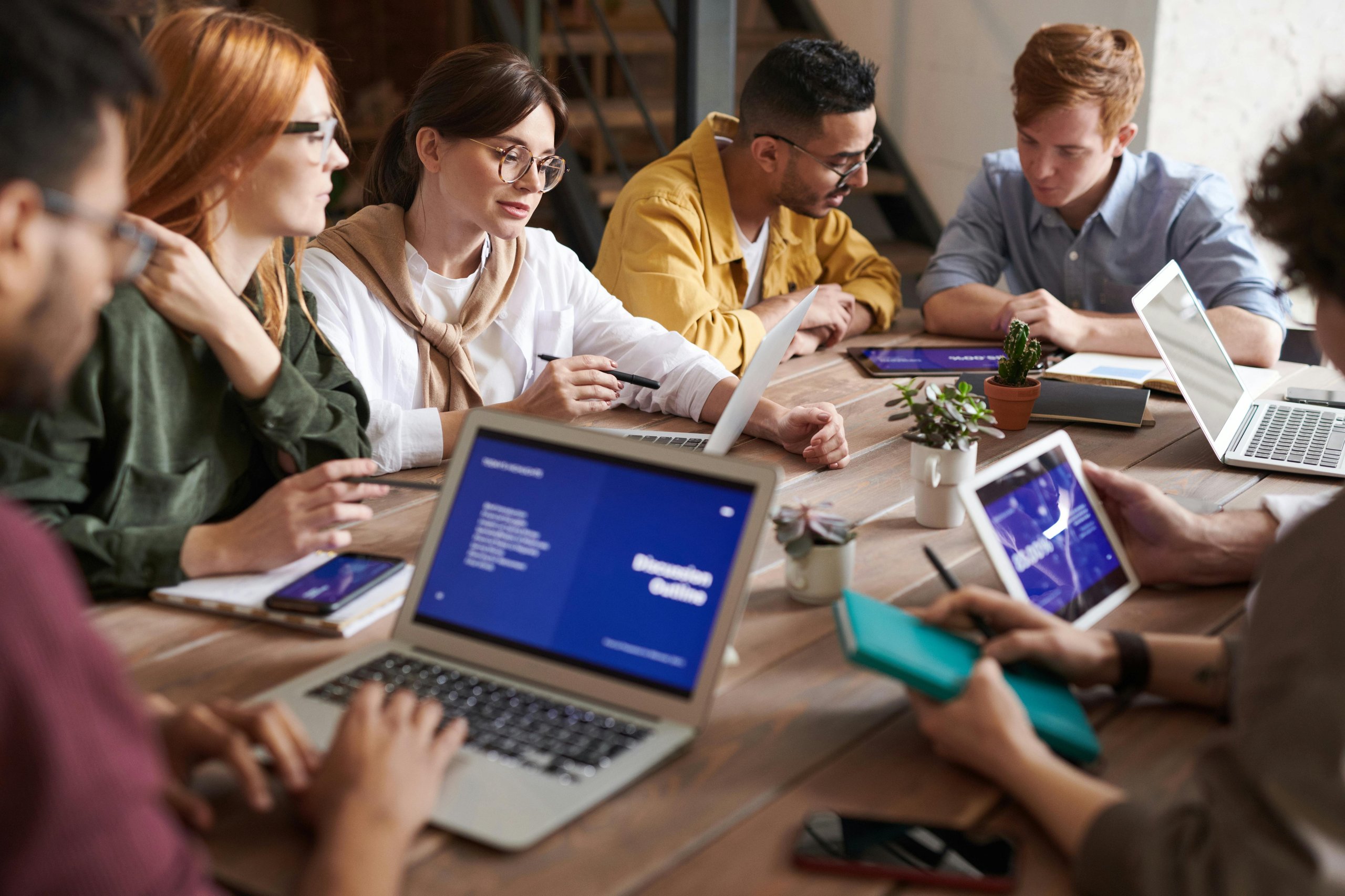 Diverse young professionals collaborating at a wooden table with laptops, tablets, and notebooks in a modern office.