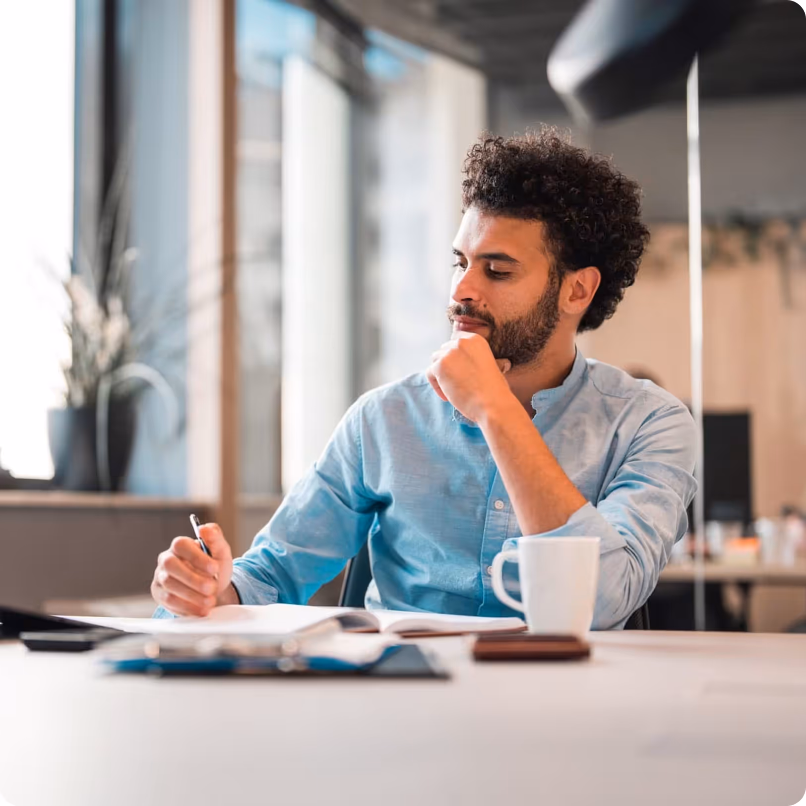 Man with curly hair and beard, wearing a light blue shirt, thoughtfully writing in a notebook at a desk with a white mug nearby in an office setting.