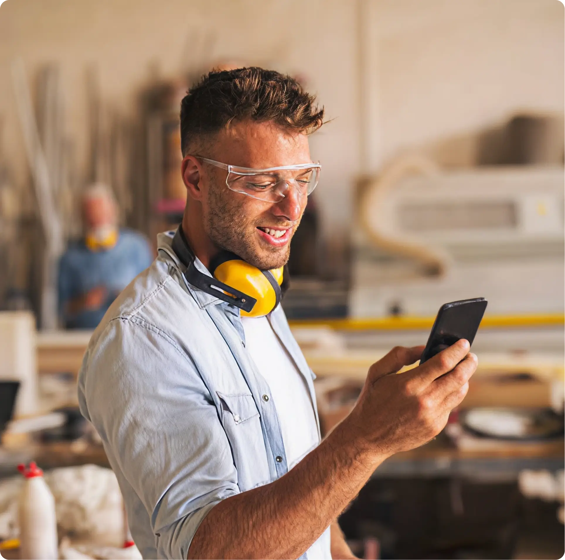 Man in safety glasses and yellow ear protection looking at his smartphone in a woodworking shop.