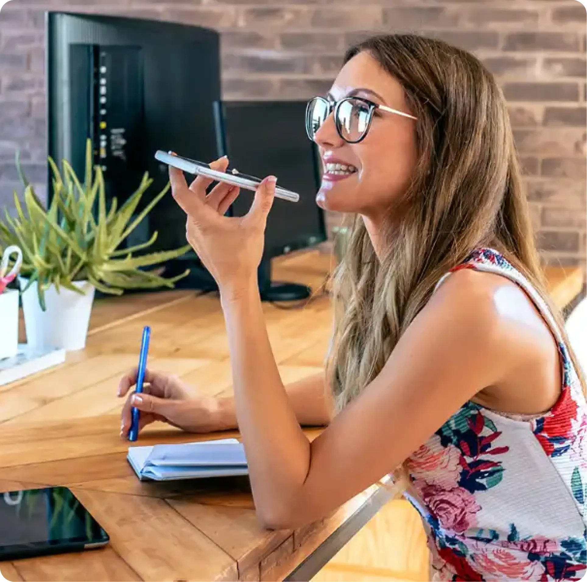 Woman with glasses smiling and holding a smartphone near her mouth while writing in a notebook at a wooden desk.