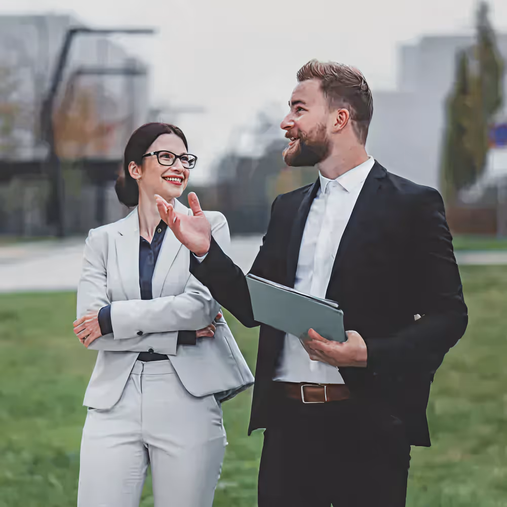 Two business professionals outdoors, a man in a black suit holding a tablet and talking, and a woman in a light gray suit smiling with arms crossed.