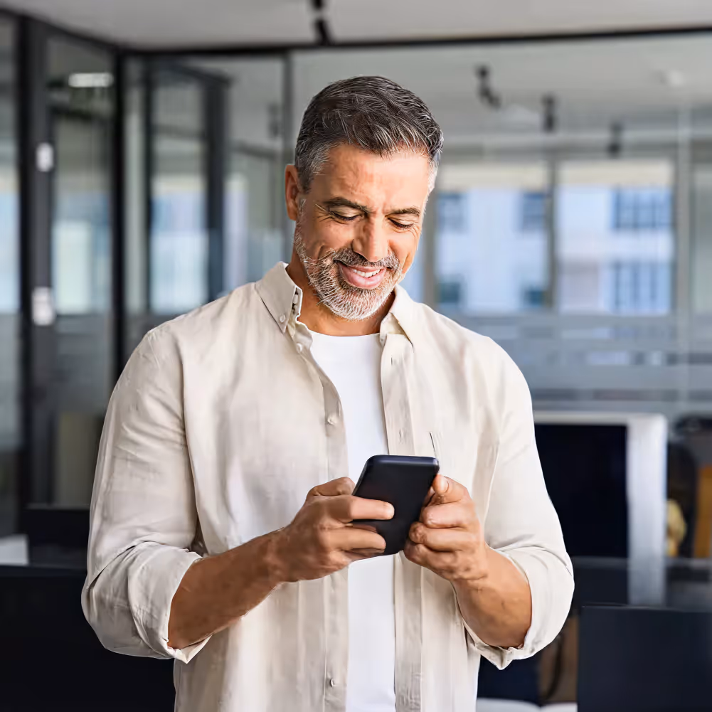Smiling middle-aged man with gray hair and beard using a smartphone in a modern office.