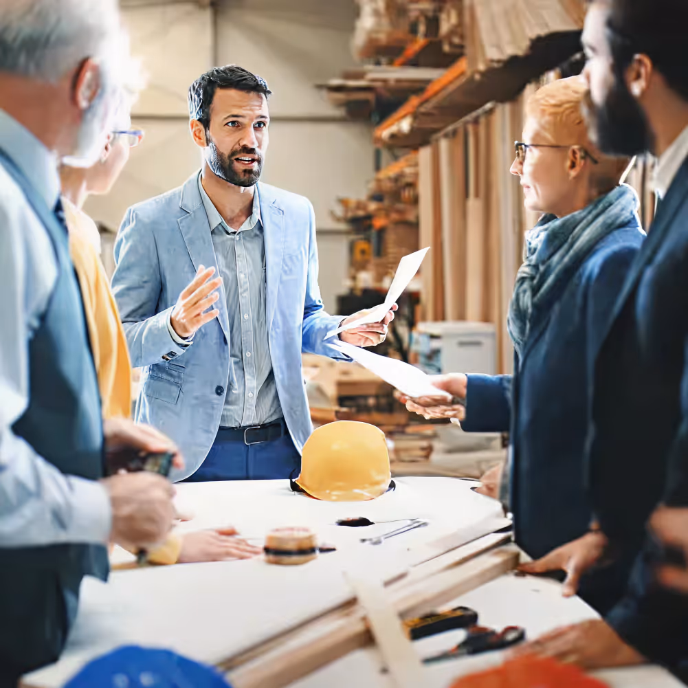 Group of professionals in a workshop discussing construction plans around a table with a yellow hard hat.
