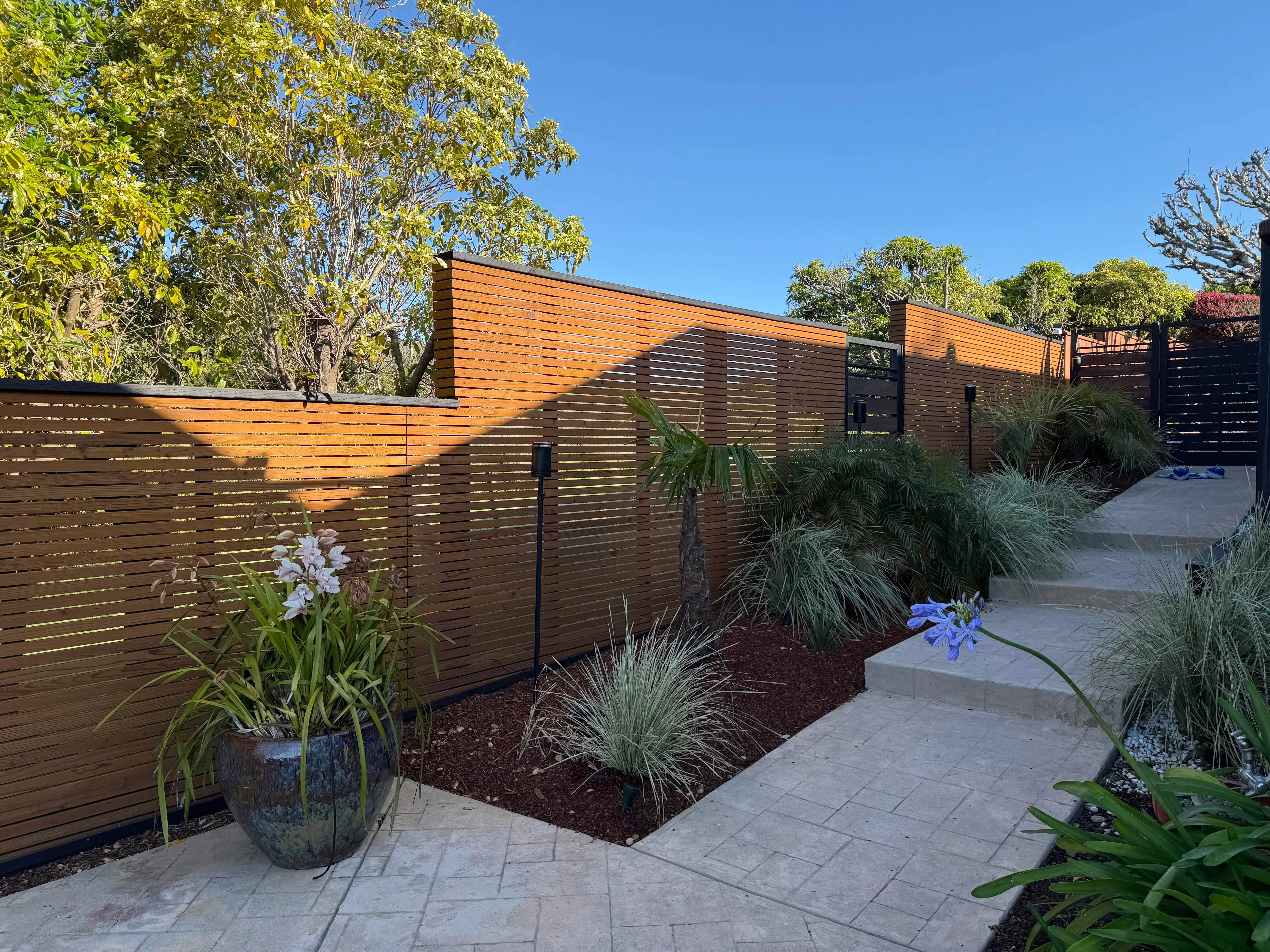 Custom horizontal wood slat fence with black posts, gate, landscaping, and paved walkway.