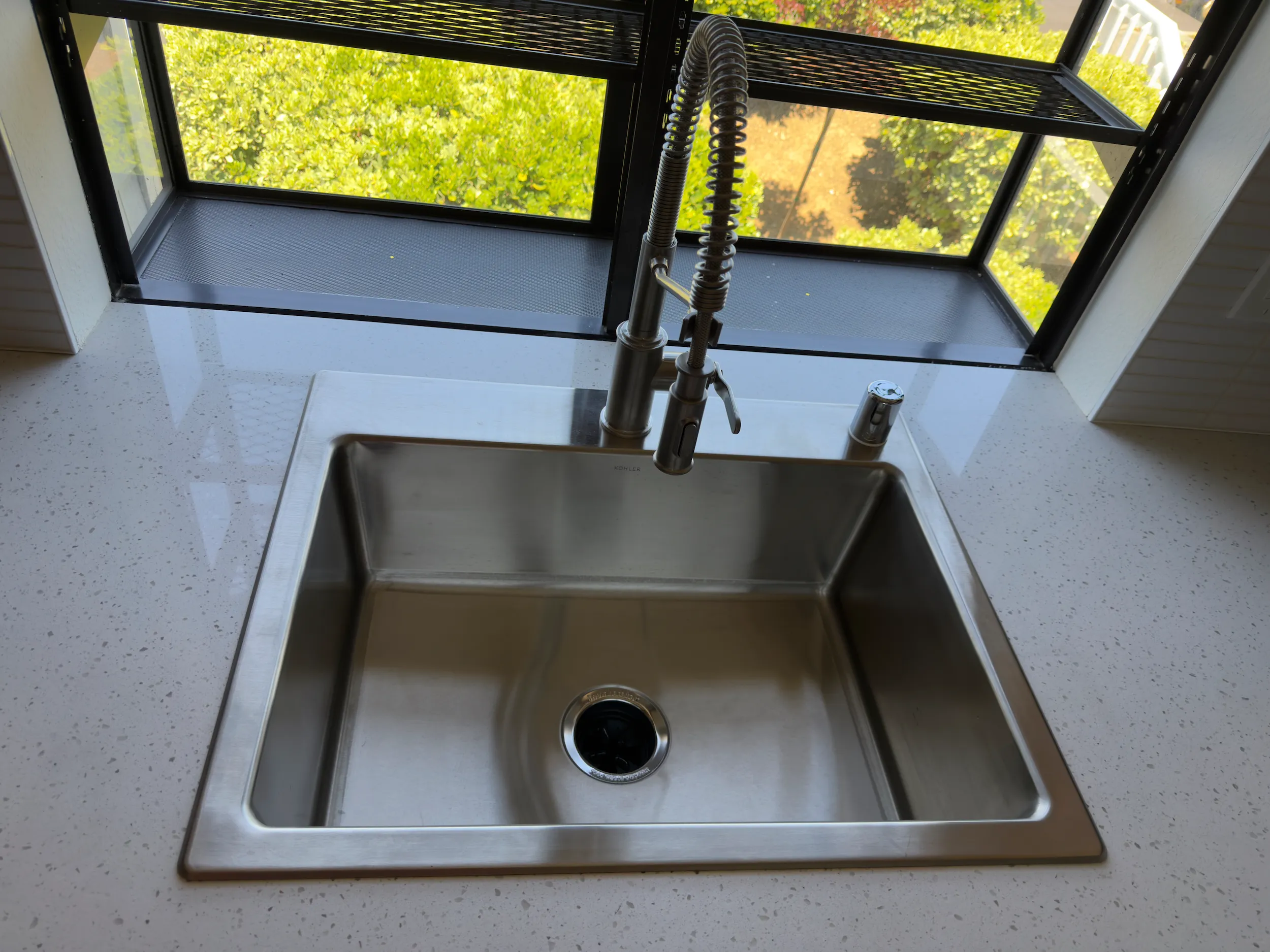 Kitchen sink and faucet installed with white countertop, window detail, and tile backsplash.