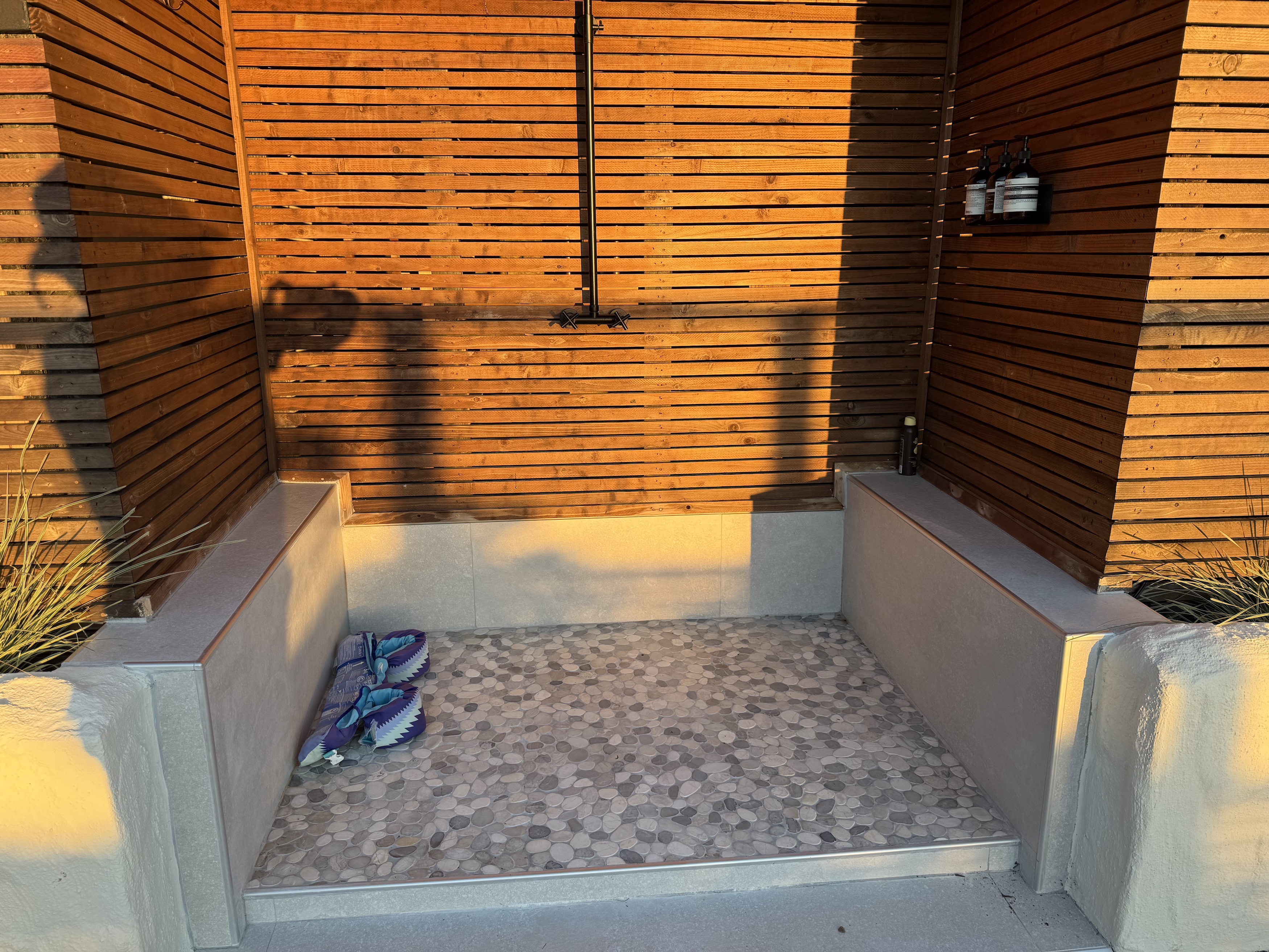 Exterior detail with custom slatted wood privacy panels, capped side walls, and pebble tile flooring at an outdoor pool shower.