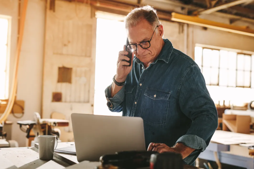 Man holding a phone while typing on a computer and standing in a workshop