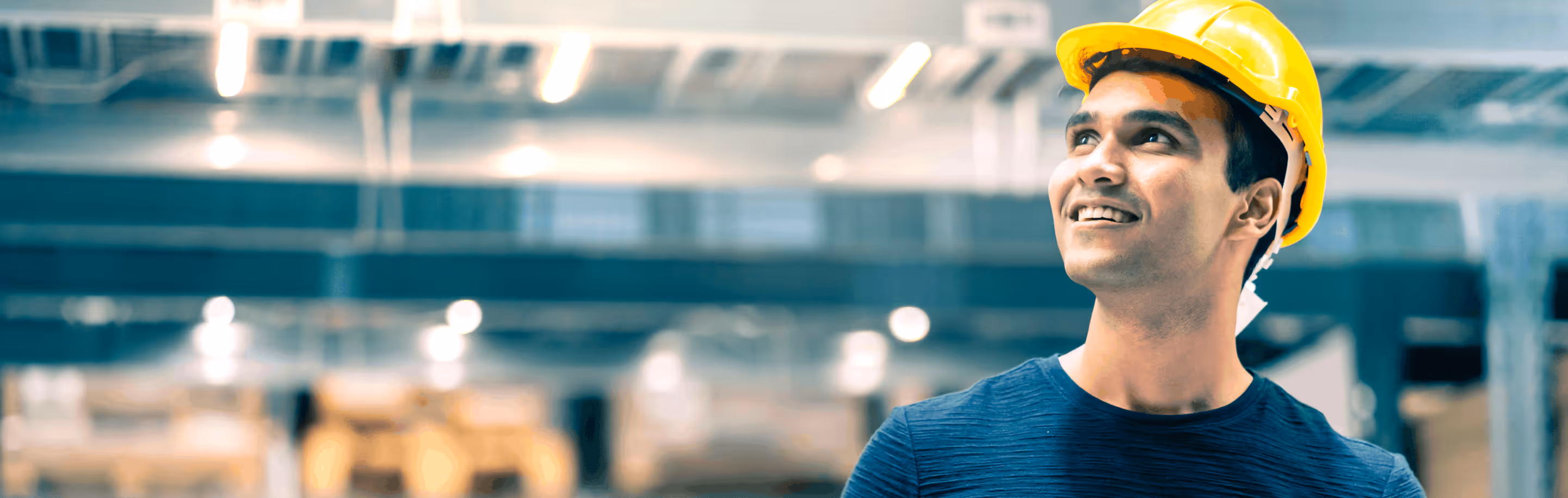 Smiling man wearing a yellow construction helmet looking upwards in a warehouse or industrial setting.