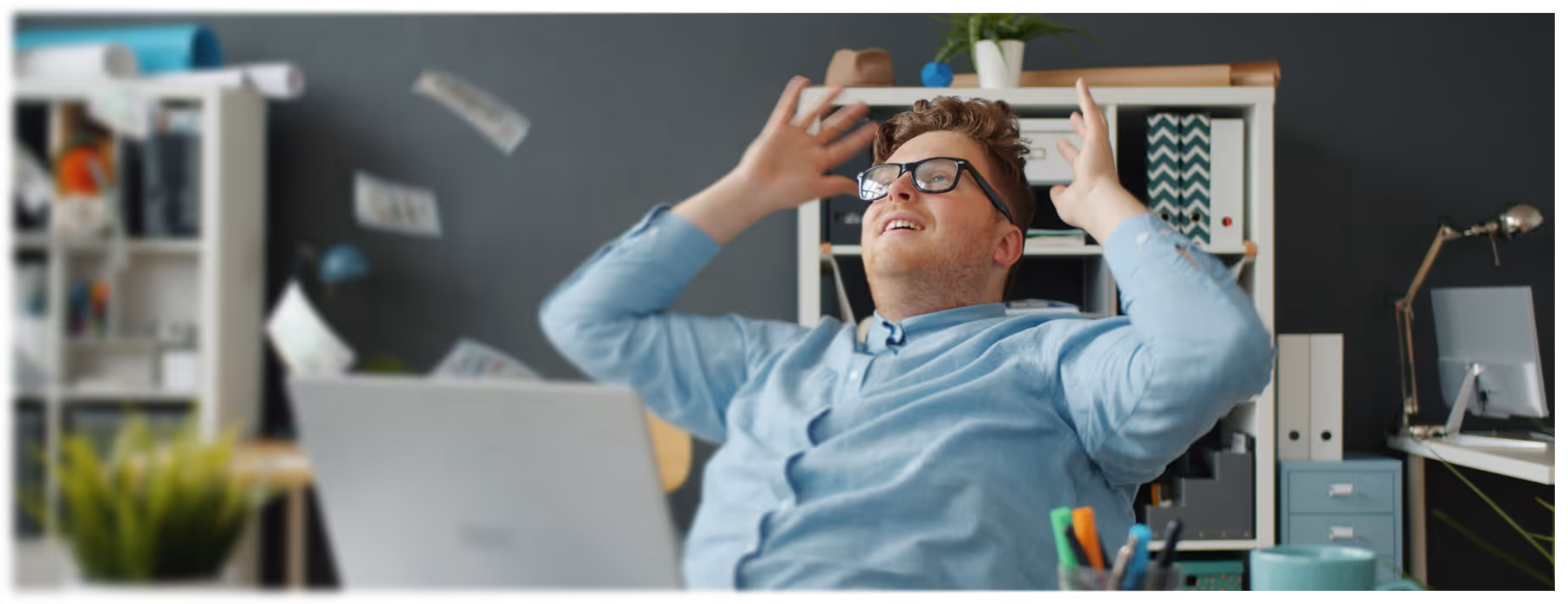 Man in glasses sitting at desk in office, throwing papers in the air with a joyful expression.