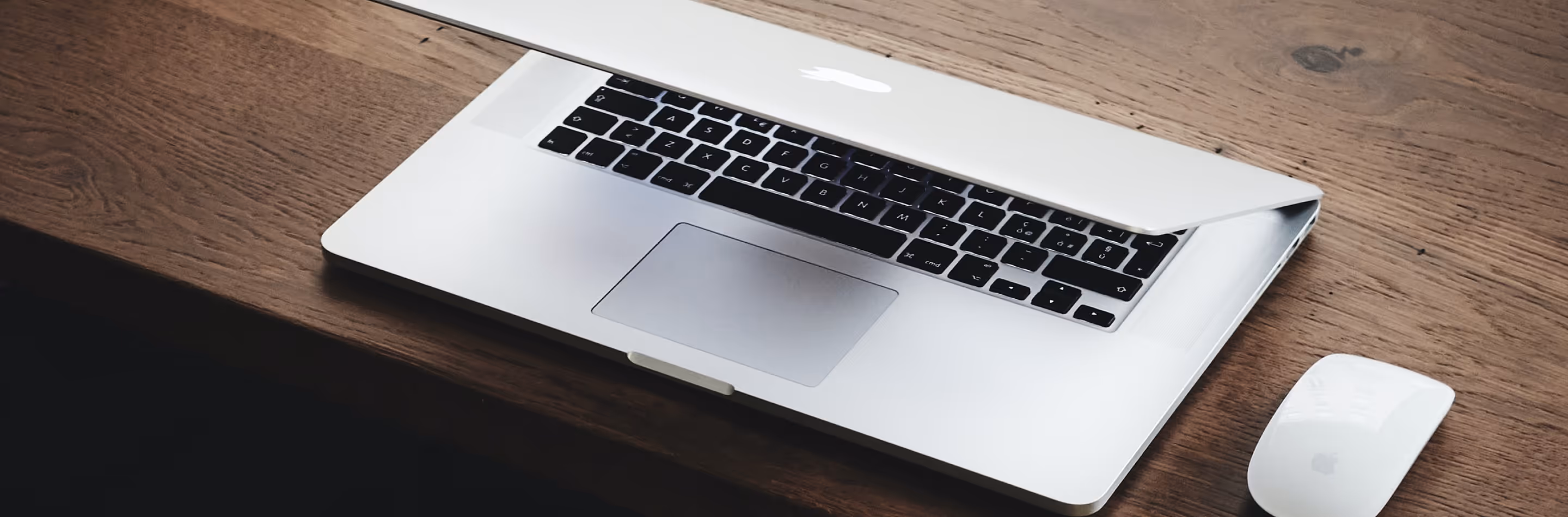 Partially closed silver laptop and white mouse on a wooden desk.