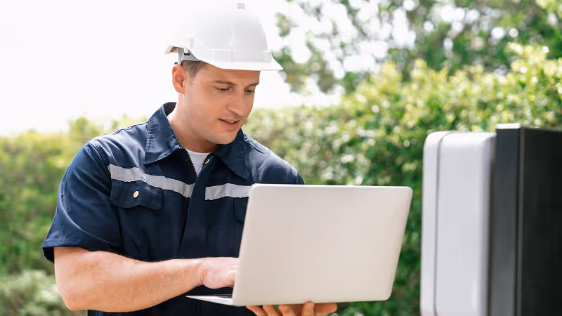 Engineer wearing a white hard hat using a laptop outdoors near electrical equipment.