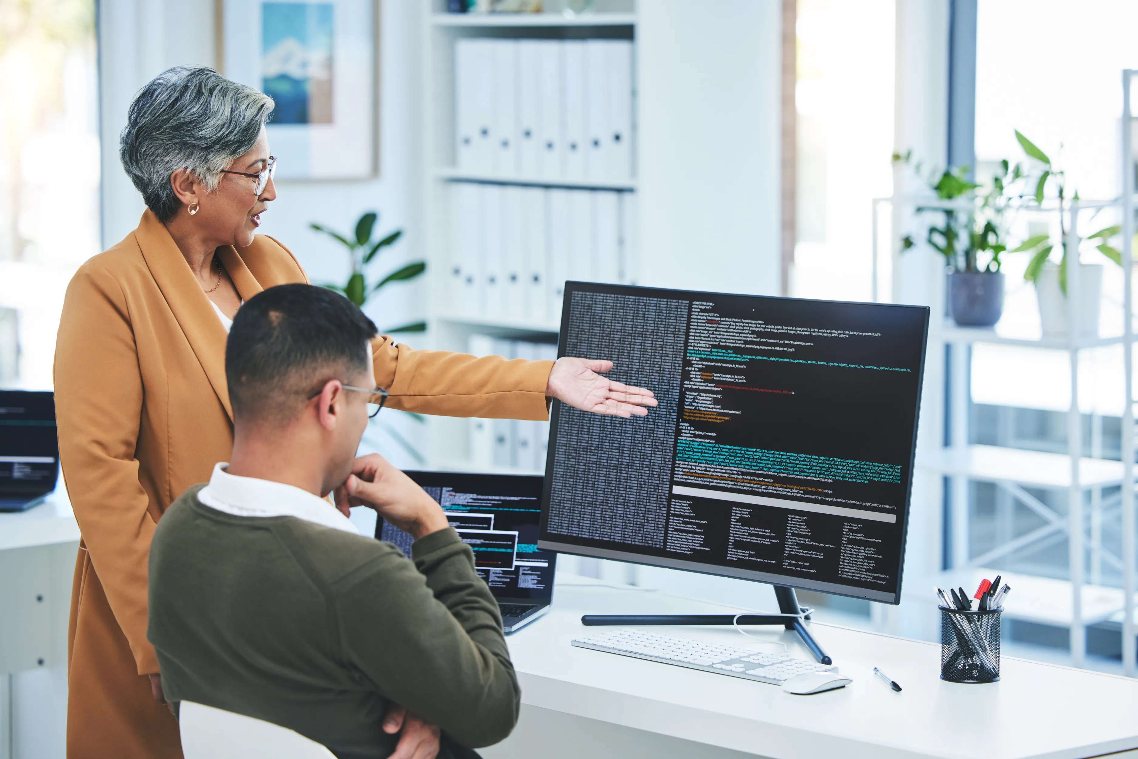 Woman in brown blazer explaining computer code on a large monitor to a seated man in an office.