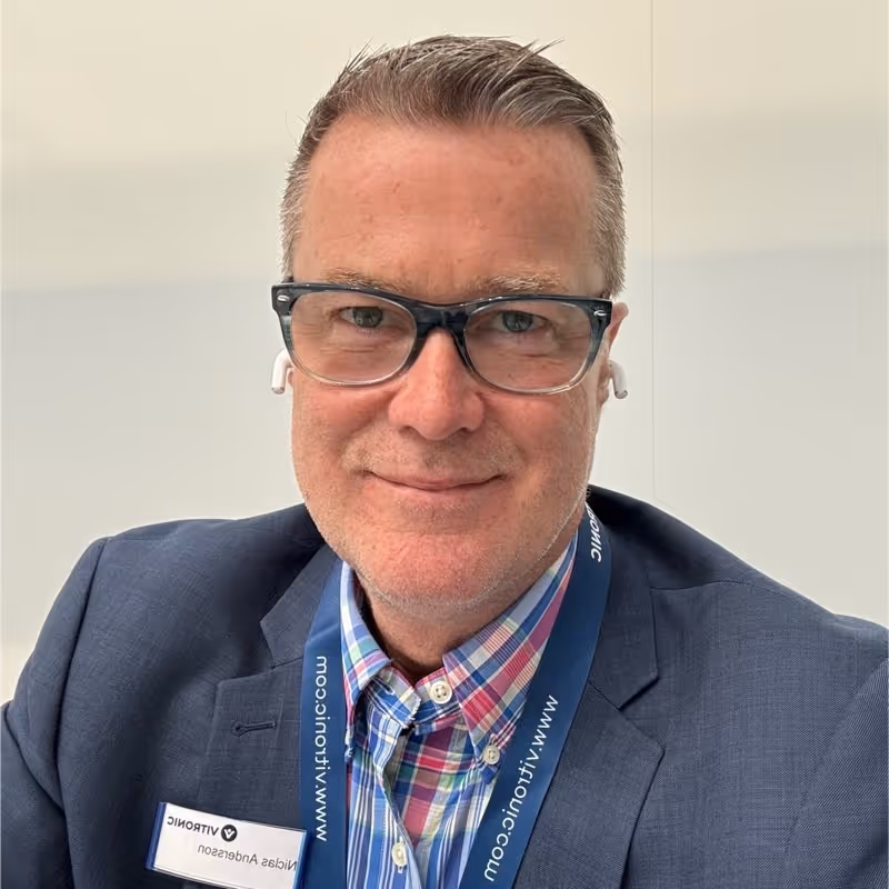 Man wearing glasses, plaid shirt, and navy blazer with a company badge and lanyard.