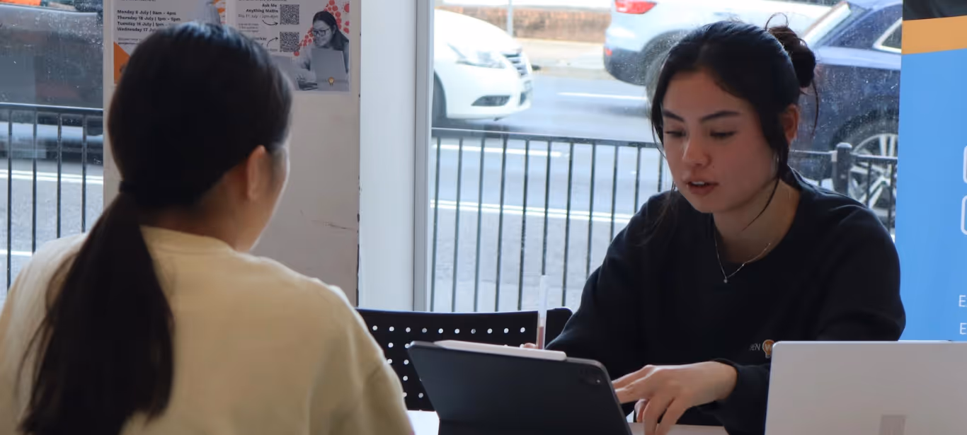 Two women talking at a table in a library with laptops and a tablet in front of them.