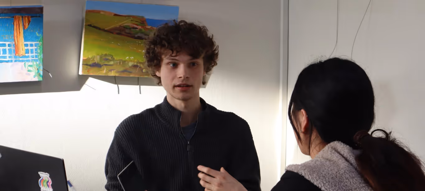 Young man with curly hair talking to a woman with a ponytail in an indoor setting with artwork on the wall.