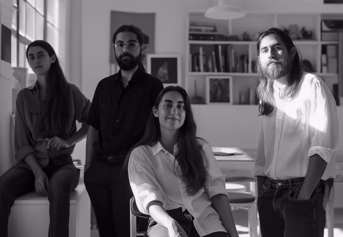 Black and white photo of four young adults with long hair posing indoors in casual clothing with a bookshelf and large window in the background.