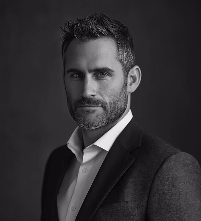 Black and white headshot of a man with short beard and styled hair wearing a dark suit jacket and white shirt, looking at the camera.