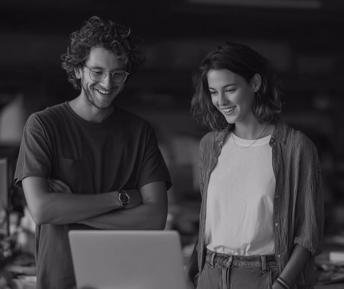 Smiling young man with glasses and curly hair standing with arms crossed and a smiling young woman with shoulder-length hair looking at a laptop together.