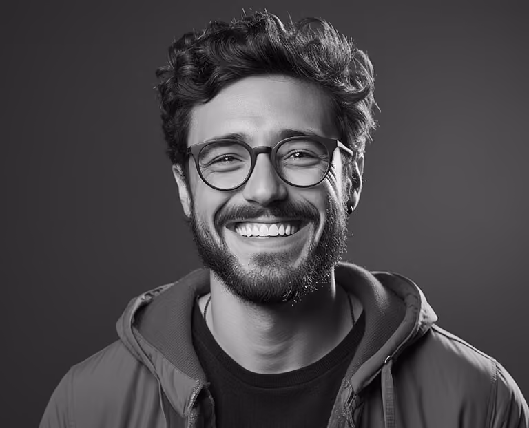 Smiling man with curly hair, beard, glasses, and a casual jacket against a plain background.