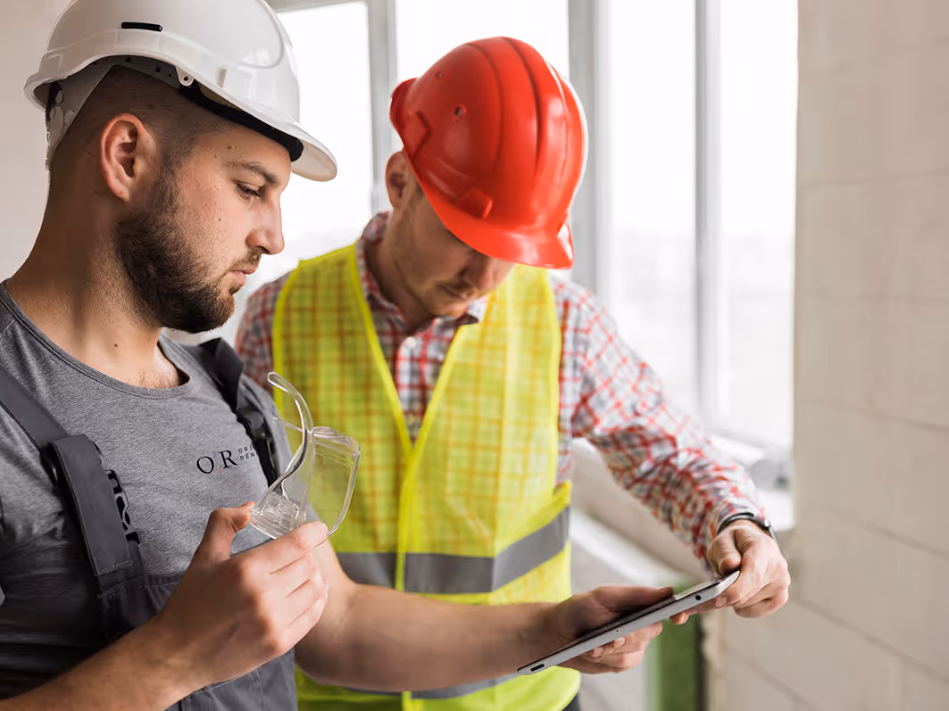 Two construction workers wearing hard hats and safety gear reviewing information on a tablet inside a building.