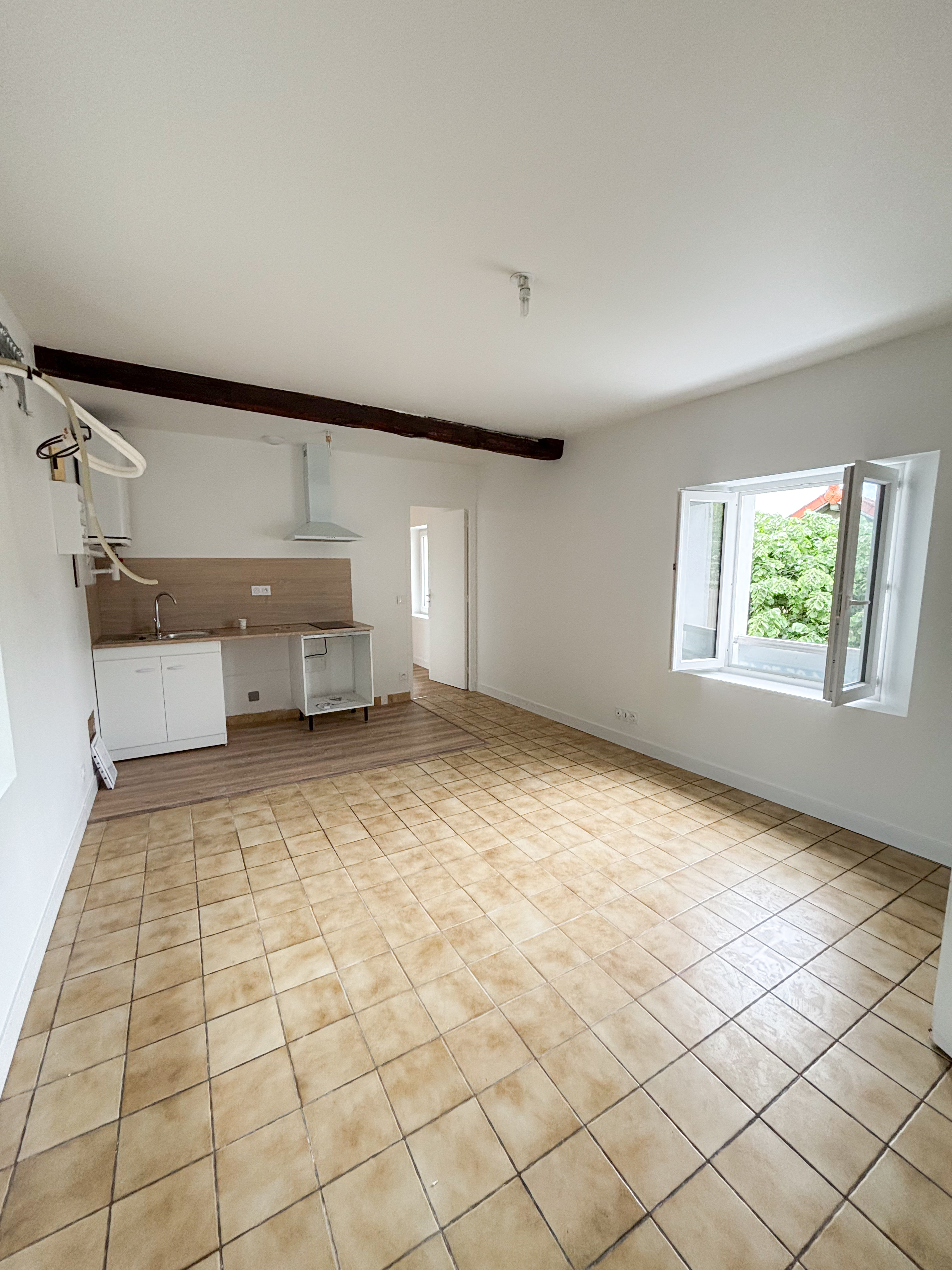 Empty room with beige tiled floor, a small kitchen area with wooden backsplash, exposed dark wooden beam on white ceiling, and an open window showing greenery outside.
