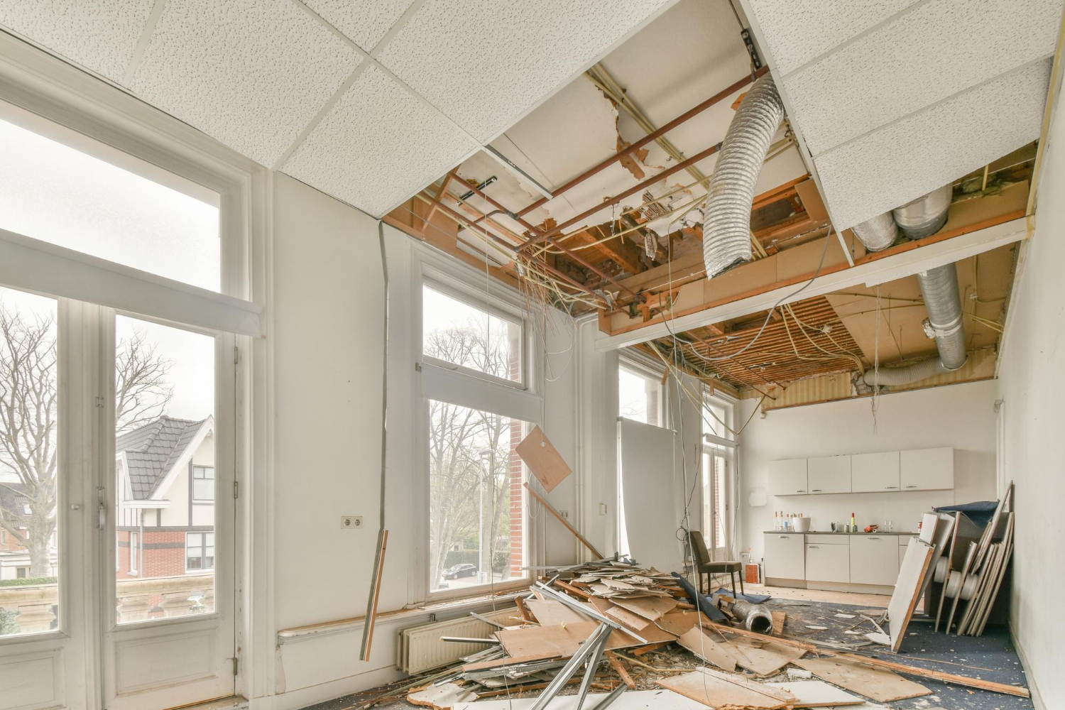 Room with large windows undergoing renovation with ceiling tiles removed, debris on the floor, and exposed pipes and wires.