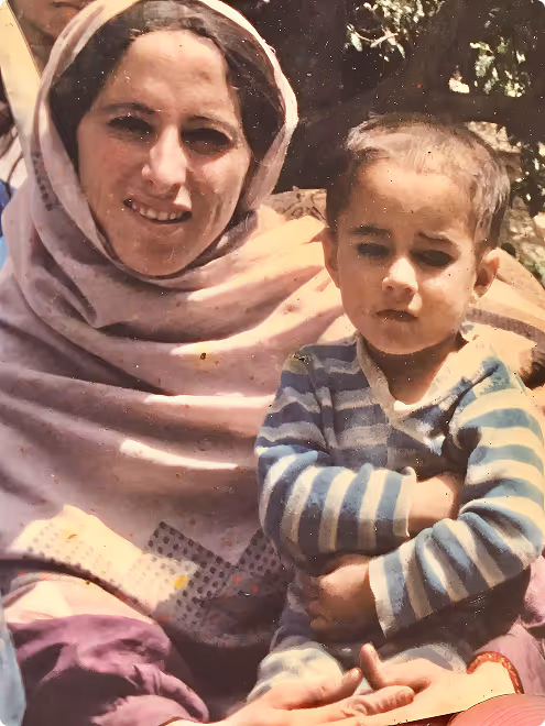 Smiling woman wearing a headscarf holding a young boy in a striped shirt with arms crossed.