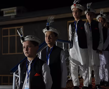 Group of boys dressed in traditional white attire with black vests and feathered hats standing in line on stairs indoors.