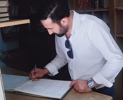 Man in white shirt leaning over a counter, writing in a large open book with sunglasses hanging from his collar.