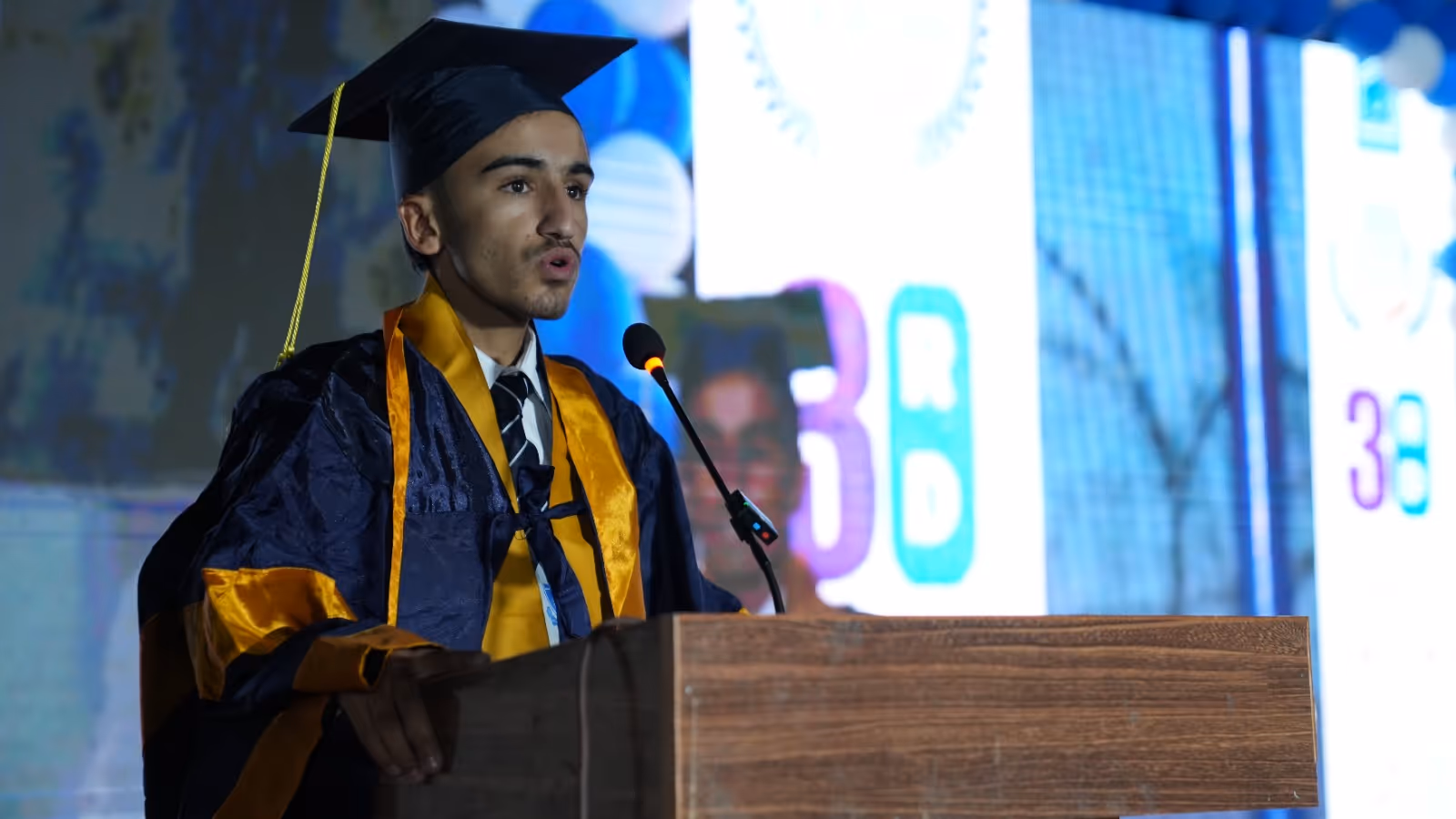 Young male graduate in cap and gown speaking at a podium with microphone during a ceremony.
