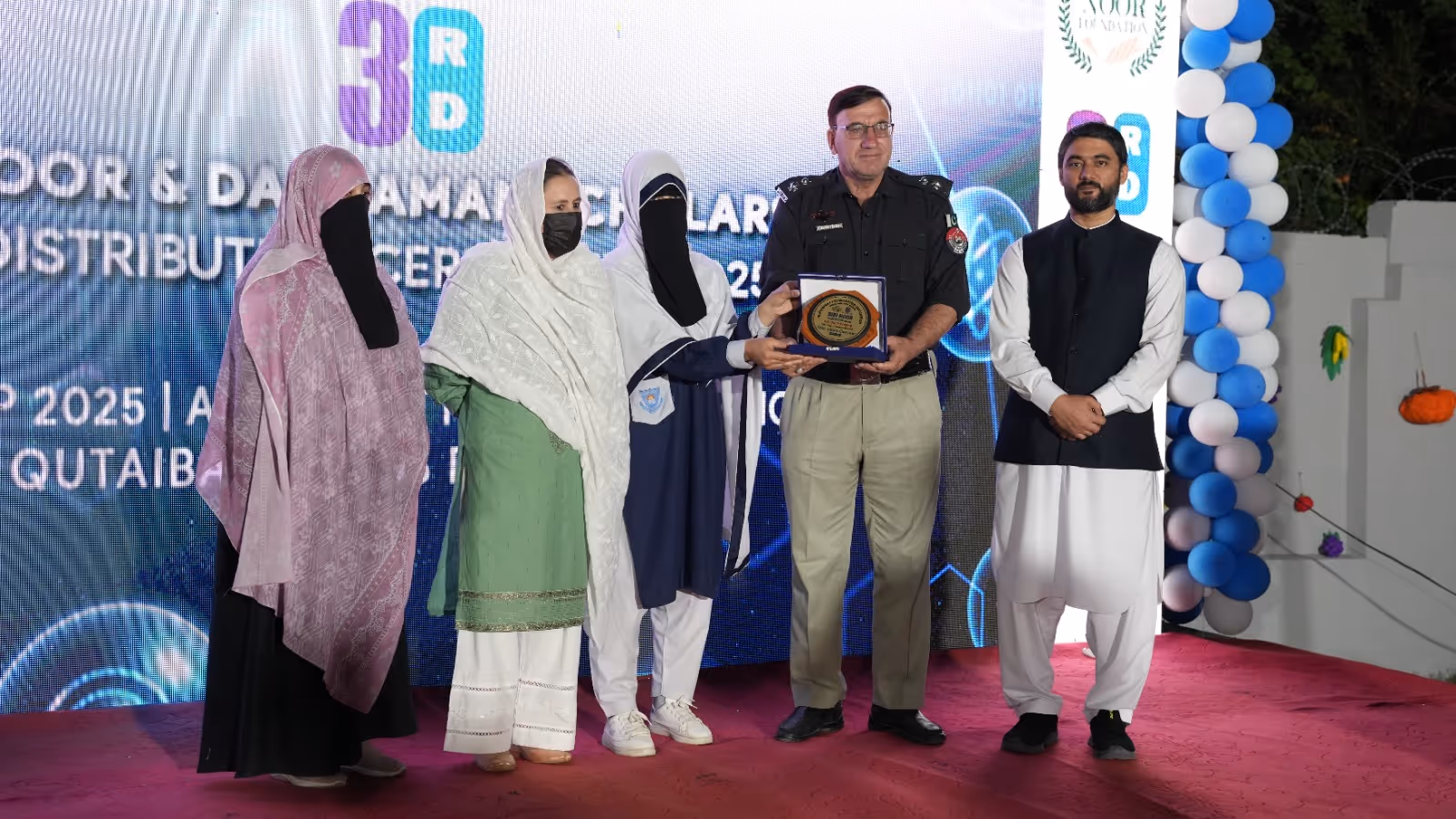 Group of five people on stage during an award distribution ceremony, with two women holding a plaque presented to a uniformed police officer.