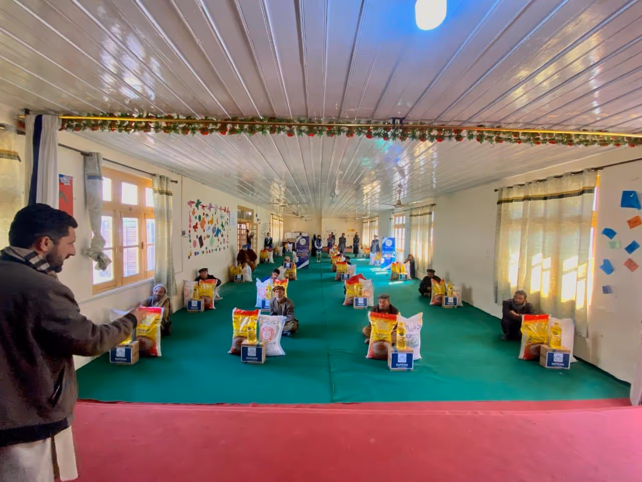 People sitting spaced apart on green carpet in a large room with bags of supplies in front of them.