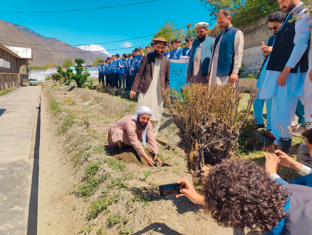 A group of men and boys outdoors, with one man planting a tree while others watch and take photos.
